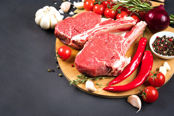Raw ribeye beef steak ready for grilling with seasonings and vegetables on wooden cutting board...