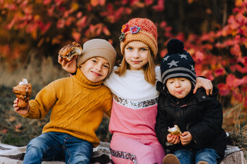 Fashionable preschool boy, handsome boy child and his smiling girl girlfriend eat a croissant, a bun, sitting on a plaid in autumn in nature. Photo of family, friendship, brother and sister, friends.