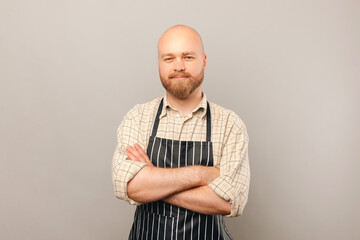 Portrait of a confident bearded barista wearing apron and holding arms crossed. Studio shot over grey background.