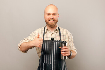 Young bald barista man holds and recommends a coffee grinder with a thumb up over light grey...