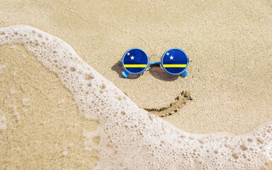 Obraz premium Sunglasses with flag of Curacao on a sandy beach. Nearby is a sea lightning and a painted smile. The concept of a successful vacation in the resorts of the Curacao.