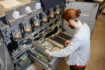 High angle portrait young man working in industrial print shop and setting up machines, copy space