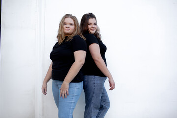 Two beautiful plump women wearing jeans and t-shirt, friends posing at the background against white street wall