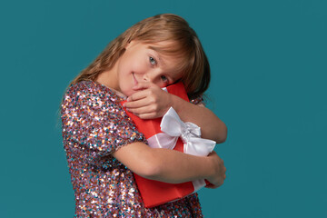 Cute cheerful child girl in a shiny elegant dress holds a red gift box in her hands on a turquoise...