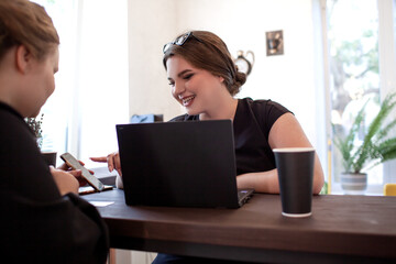 Two overweight business women sit in cafe with cup of coffee or tea and looking at the laptop, plus size female is holding a bank card to place an online order shopping