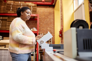 Side view portrait of adult black woman operating industrial printing machine in workshop, copy...