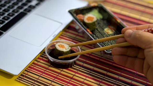 Close up, male hand picks up a piece of sushi with chopsticks, next to a laptop.