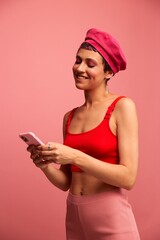 Naklejka premium Young woman blogger with colored pink hair and short haircut flipping through the screen of the phone and typing a message with a smile in stylish clothes on a pink background
