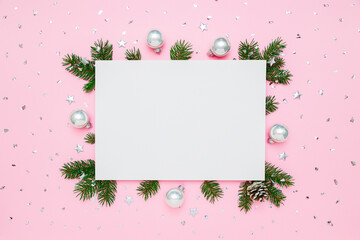 Christmas, New Year holiday card with copy space for text. Blank white paper in frame of natural fir tree branches and silver decorations on pastel pink flat lay background