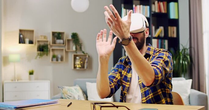 Caucasian man with beard in VR headset sitting at table and having virtual reality experience at home. Inside. Male user and gamer playing and moving hands in air. Technology of metaverse.