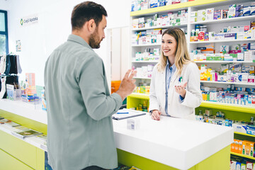 Customer entered in pharmacy store and looking for some medical pills and cremes. Young pharmacist woman is helping him