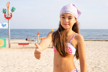 Close-up Portrait of a pretty little girl with long hair with thumb up standing on the beach