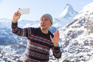 Adult man in warm knitted sweater and hat traveling in winter Swiss Alps, recording video with...