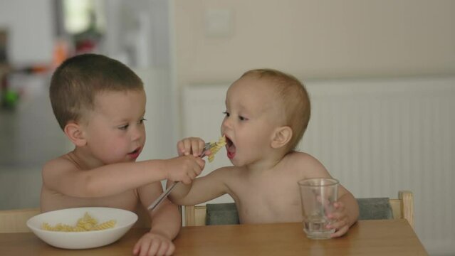 Two Little Boys Feed Each Other Sitting At Table In Kitchen. Caring Friends Kids. Siblings Brothers Eating Together, Elder Baby Child Feeds Younger Brother Pasta Food On Fork, Putting Bite Into Mouth