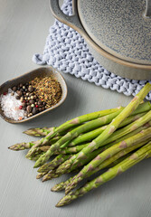 Closeup view of a bunch of green asparagus next to a tray full of spices and a ceramic pot over a blue knitting cloth.