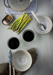Ingredients for an asparagus soup dinner scene with green asparagus, two ceramic bowls and a ceramic pot over a wooden gray table.