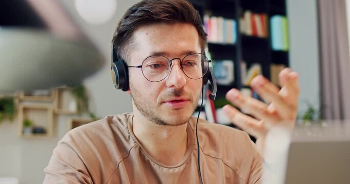 Close up of Caucasian male vlogger in glasses and headset with mic talking to webcam of laptop. At home. Young man having meeting online and consulting client while working. Indoor.