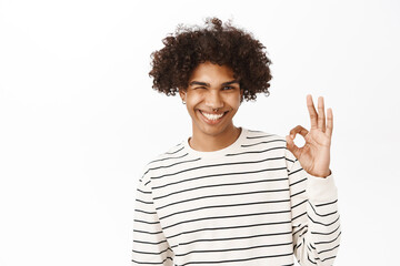 Close up portrait of smiling hispanic guy shows okay, ok sign, approves smth, recommends, stands over white studio background