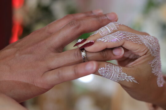 A picture of a bride putting a wedding ring on the groom's hands with white henna tattoo, red fake nails and blurry background 