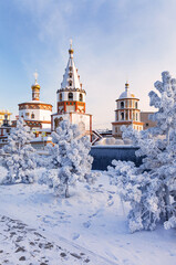  Irkutsk on frosty winter day. View from the Lower Embankment of the Angara to the beautiful Epiphany Cathedral in the Baroque style. Winter city landscape with snow-covered fir trees in the park