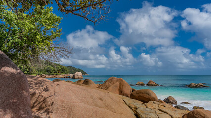 Piles of picturesque granite boulders on the shore of the turquoise ocean. A green hill in the distance. Tree branches against a background of blue sky and clouds. Seychelles. Praslin. 
Anse Lazio