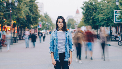 portrait of tired young woman student standing alone in city center and looking at camera with straight face while crowds of men and women are whizzing around. - 542858945