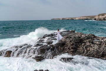Fototapeta na wymiar A woman stands on a rock in the sea during a storm. Dressed in a white long dress, the waves break on the rocks and white spray rises.