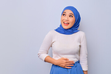 Satisfied young Asian Muslim woman keeps both hands on belly, after delicious dinner isolated over white background