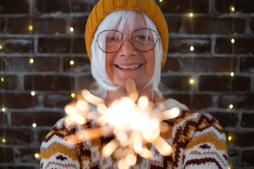 Handsome senior caucasian woman with eyeglasses and winter sweater holding sparkler light, elderly...