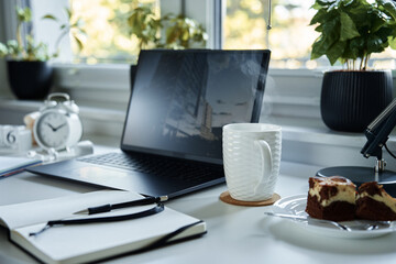 Home office workplace. Table with laptop, notebook and hot coffee near window in living room.