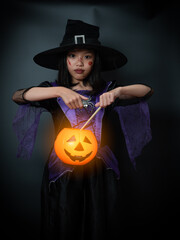 Little girl cosplay as a witch and makeup on a celebration of Halloween. Child in costume and holding pumpkin bucket in dark background.