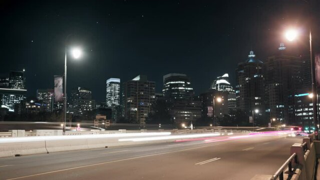 Night timelapse of traffic on the bridge, with cityscape in the background and starry sky. Zoom out. 
