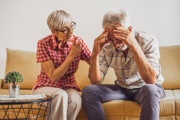 Senior couple sitting in living room. Man is sad and worried and woman is consoling him.