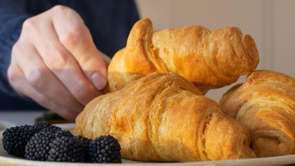 Close-up of fresh croissants, one which is taken by man and eats. Delicious and crispy croissants fresh from bakery for delicious breakfast. Blackberries and freshly squeezed juice complete croissants