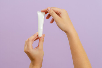Hand holds plastic white tube isolated on lilac background. Beauty concept. Packaging tube for cosmetic products