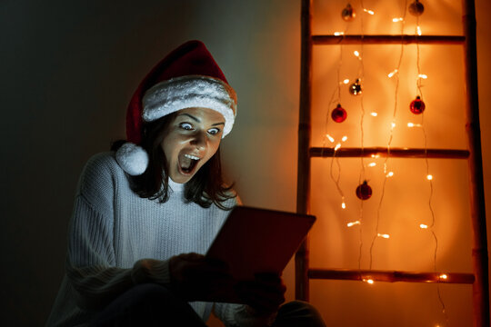 Woman in Santa hat holding digital tablet in her hands looking at screen and screaming with happiness.