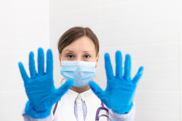 Beautiful young doctor woman in a medical mask and gloves with a stethoscope in the hospital shows hand gestures. Selective focus. Portrait. Close-up