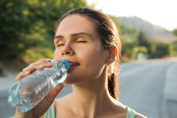 Happy young woman drinking water outdoors on hot summer day. Refreshing drink