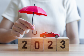 hand holding umbrella and cover red car toy with 2023 Year block on table. Car insurance, warranty, repair, Financial, banking, money and New Year concept