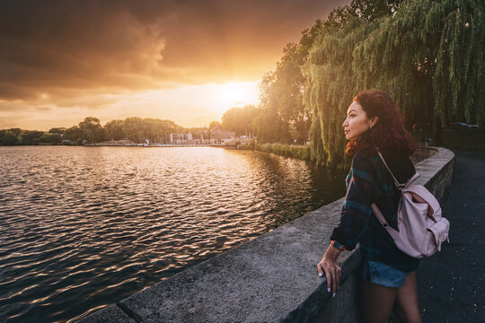 A student girl with a backpack stands near the embankment of Lake Aasee in Munster and enjoys a stunning sunset view