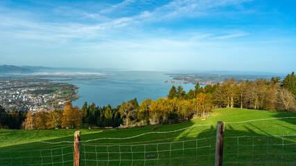 Fototapeta premium Herbstliches Panorama vom Pfänder über den Bodensee, Dunst und Saharasand liegen in der Luft und schaffen ein mystisches Bild mit orangen und gelben Laubbäumen, Bregenz, Stadt im Tal. Bodensee
