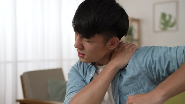 tired korean male manager having a sudden crick in neck is exercising stiff shoulder muscles and pounding massaging himself while working from home