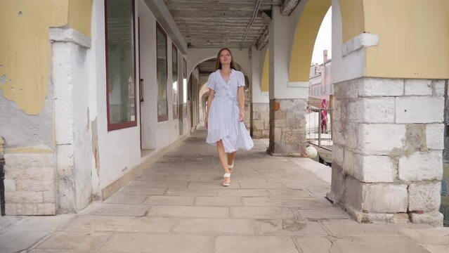 Young and beautiful woman walks under Venetian arcades near the canal with boats