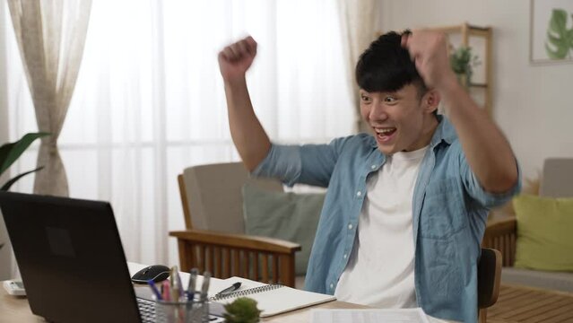 excited asian male working from home is screaming with clenched fists feeling happy about getting job promotion online notification on the computer