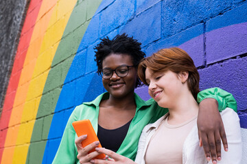 Smiling woman sharing smart phone with friend in front of rainbow colored wall