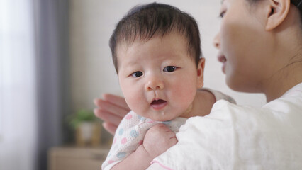 close up shot adorable baby girl in her mom’s arms is getting little upset while being burped and patted on the back at home.