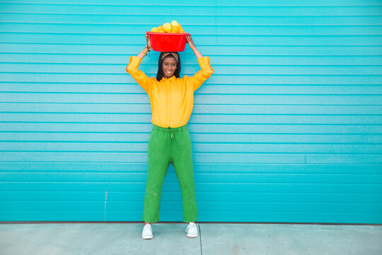 Woman carrying basket full of oranges on head in front of purple wall