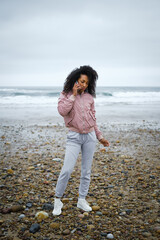 Young fashionable sporty afro hairstyle woman talking on the phone at the beach. Autumn season sportswear fashion.