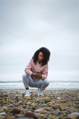 Young fashionable sporty afro hairstyle woman texting on smartphone at the beach. Autumn season sportswear fashion.