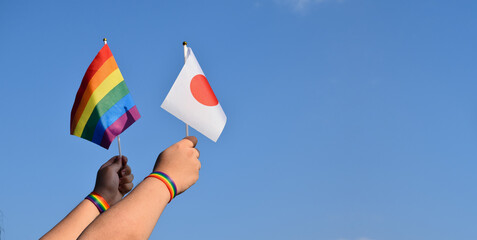 Japan flag and rainbow flag raising in hands which has rainbow wristband and against bluesky, soft...